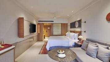 A modern hotel room with a bed, a sofa, a dining table, and a TV on the wall.
