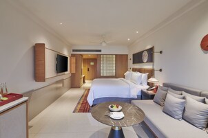 A modern hotel room with a bed, a sofa, a dining table, and a TV on the wall.