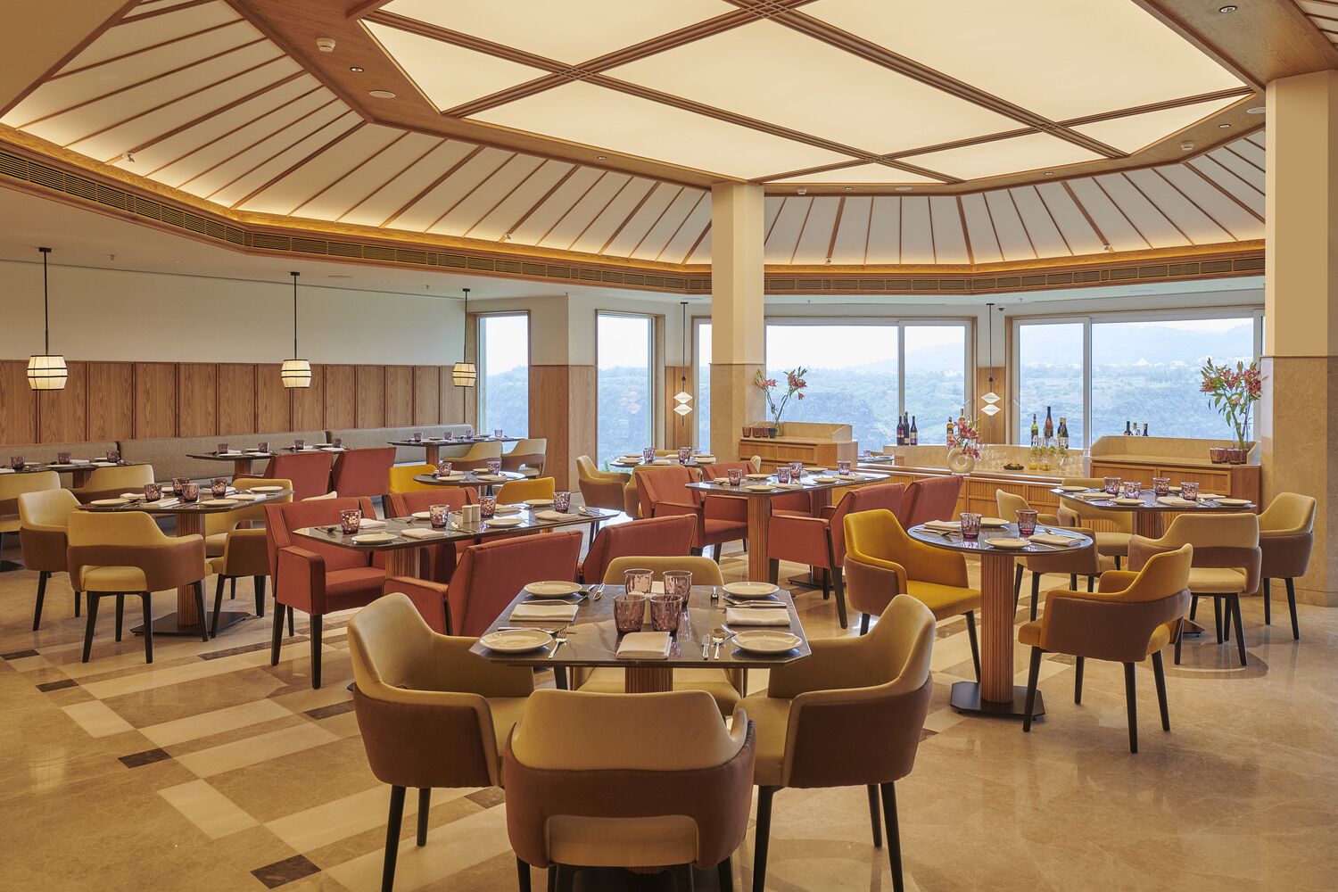 Un restaurante espacioso con techos altos y ventanas amplias que ofrecen vistas a la montaña. Las mesas están dispuestas con platos y cubiertos, y el suelo está cubierto de baldosas.