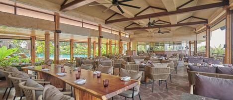 A spacious outdoor dining area with multiple tables and chairs, large windows, and ceiling fans.