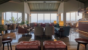A spacious lobby with a high ceiling, large windows offering a view of the mountains, and a bar area with shelves of bottles. - Athiva Resort & Spa, Khandala (Mawal)