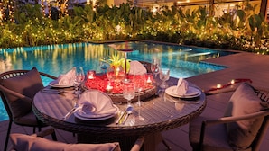 A dining table set for two with a pool view, surrounded by wicker chairs. - Athiva Resort & Spa, Khandala (Mawal)