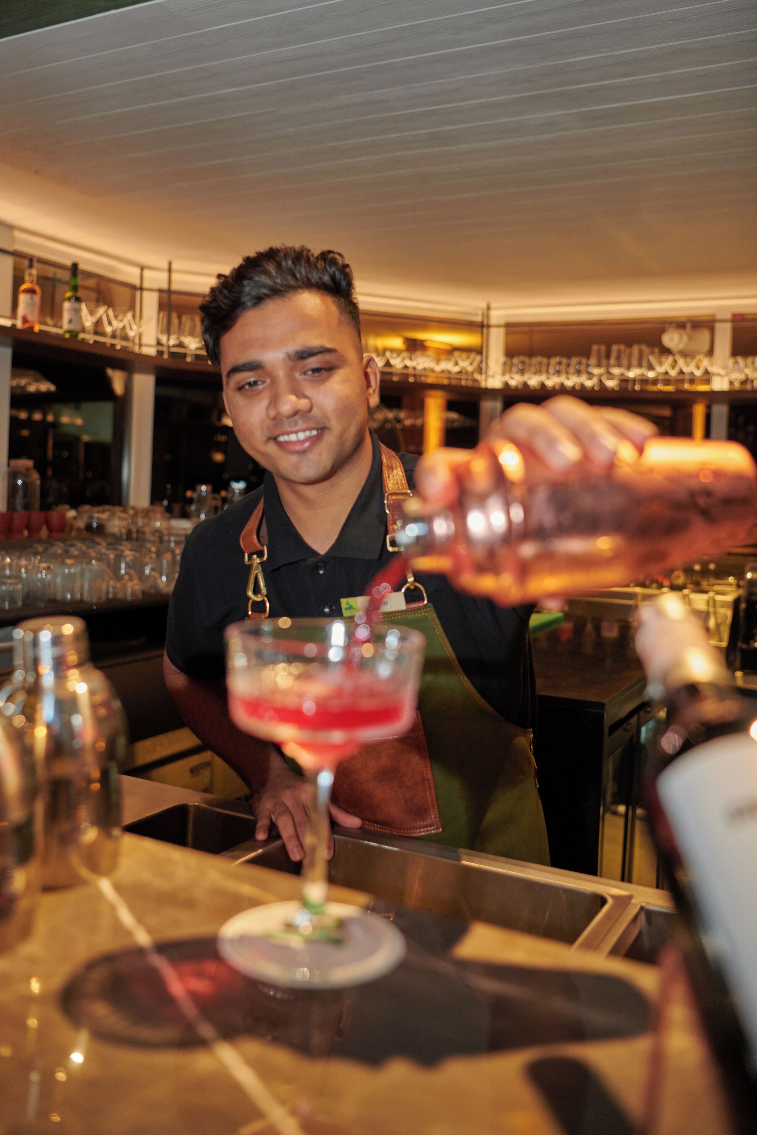 En bartender forbereder en drink bag en bar, med hylder fyldt med flasker og glas i baggrunden.