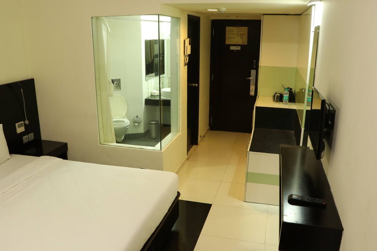 A hotel room with a bed, a bathroom with a glass shower enclosure, a desk with a computer, and a TV.