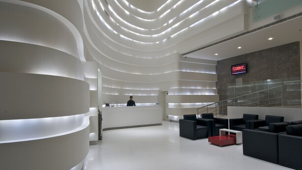 A modern reception area with a curved white reception desk, a digital display, and a staircase leading to upper levels. - Keys Select by Lemon Tree Hotels, Thiruvananthapuram (Thiruvananthapuram)