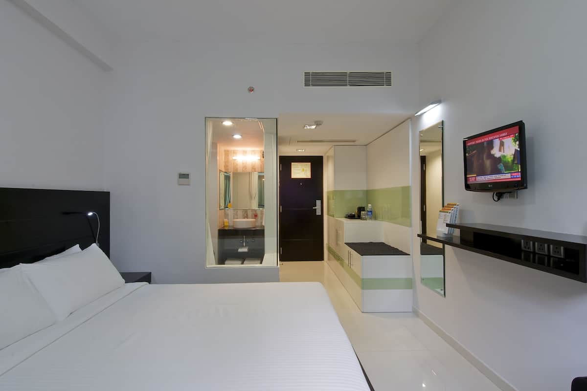 A modern hotel room with a bed, a TV mounted on the wall, a small desk, and a bathroom visible through a glass door.