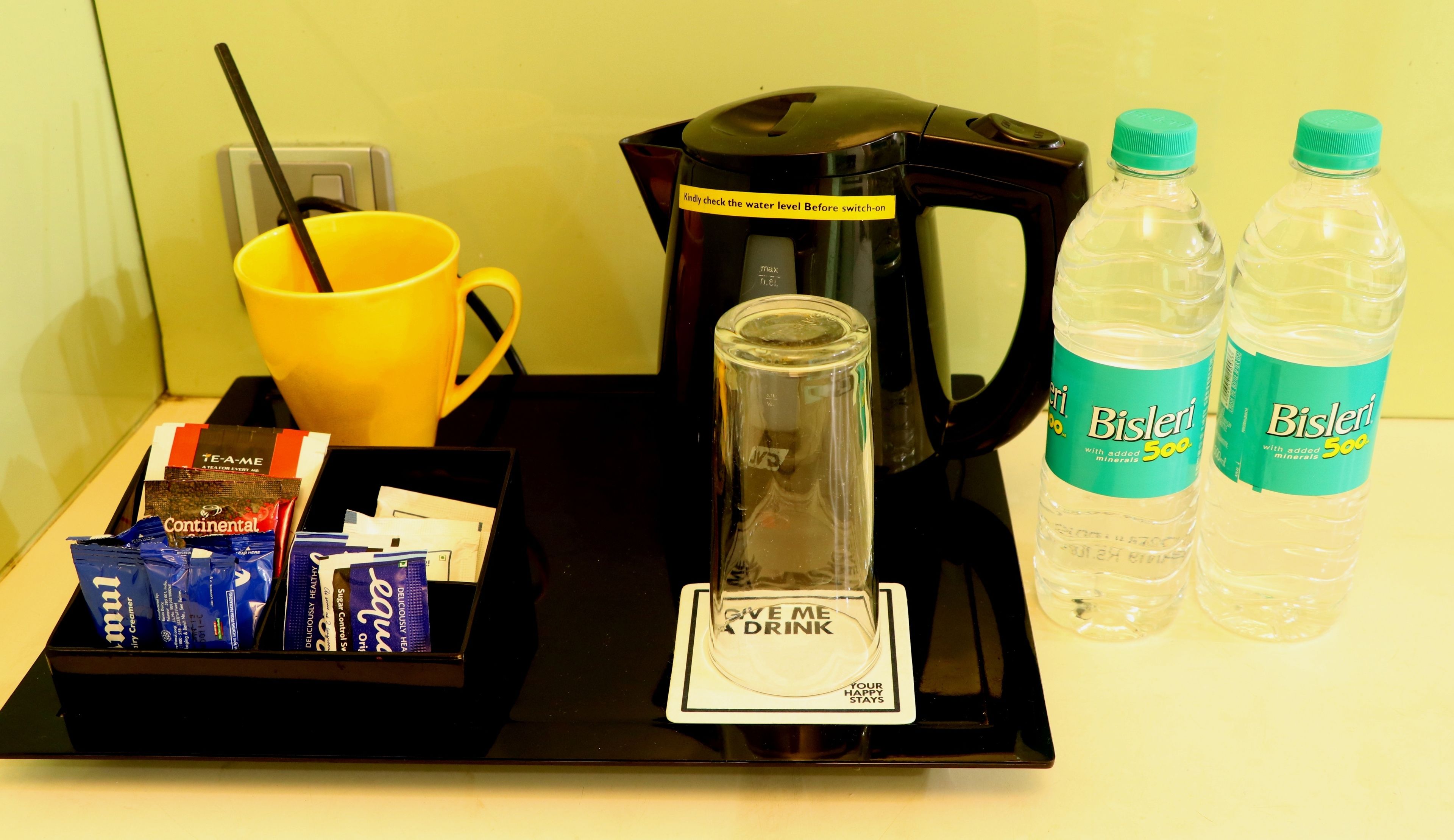 Una cafetera, una taza amarilla, un vaso con posavasos y dos botellas de agua Bisleri sobre una bandeja.