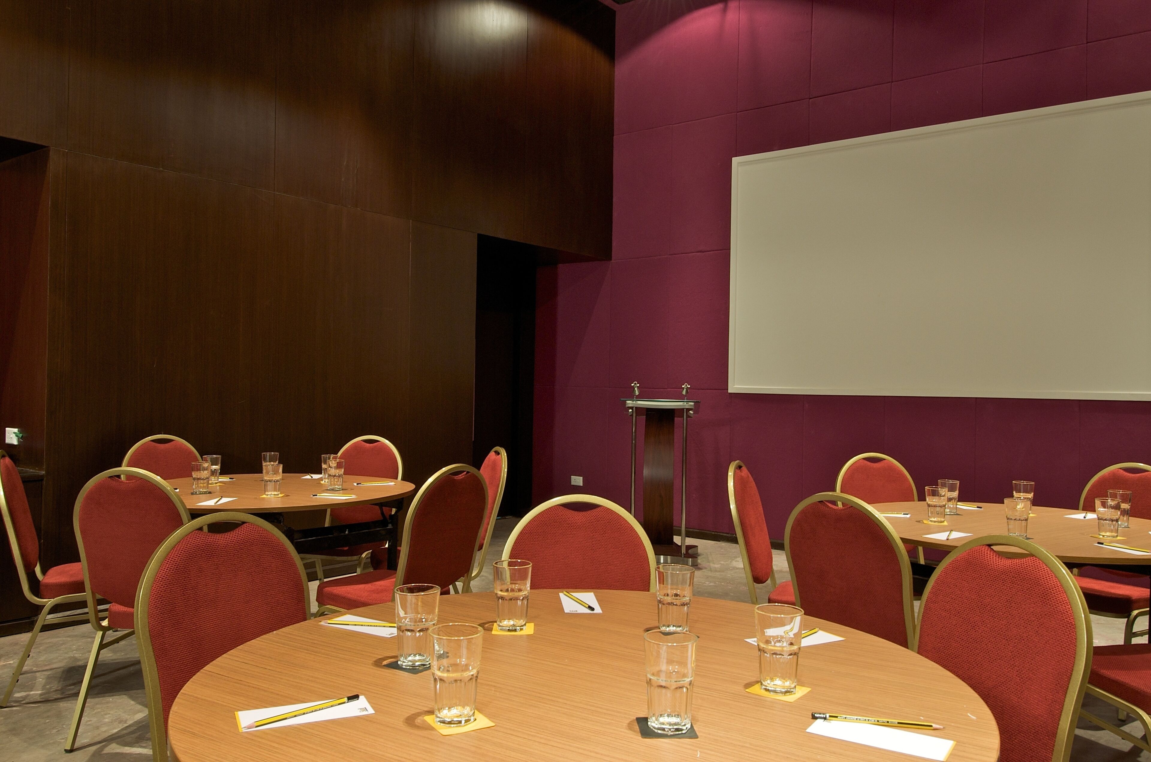 Une salle de conférence avec des tables rondes, des chaises rouges et un écran de projection.