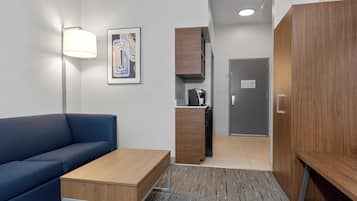 A modern hotel room with a blue sofa, a wooden coffee table, a desk, a kitchenette, and a wardrobe.