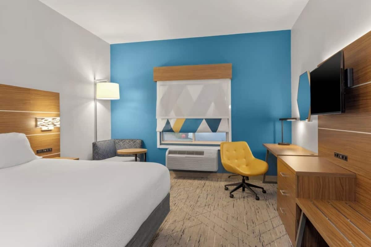 A hotel room with a bed, a desk, a chair, a TV, and a window with a geometric curtain.