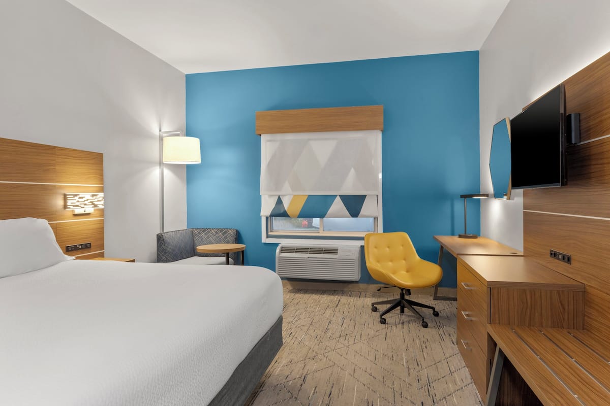 A hotel room with a large bed, a desk, a chair, a TV, and a window with blinds.