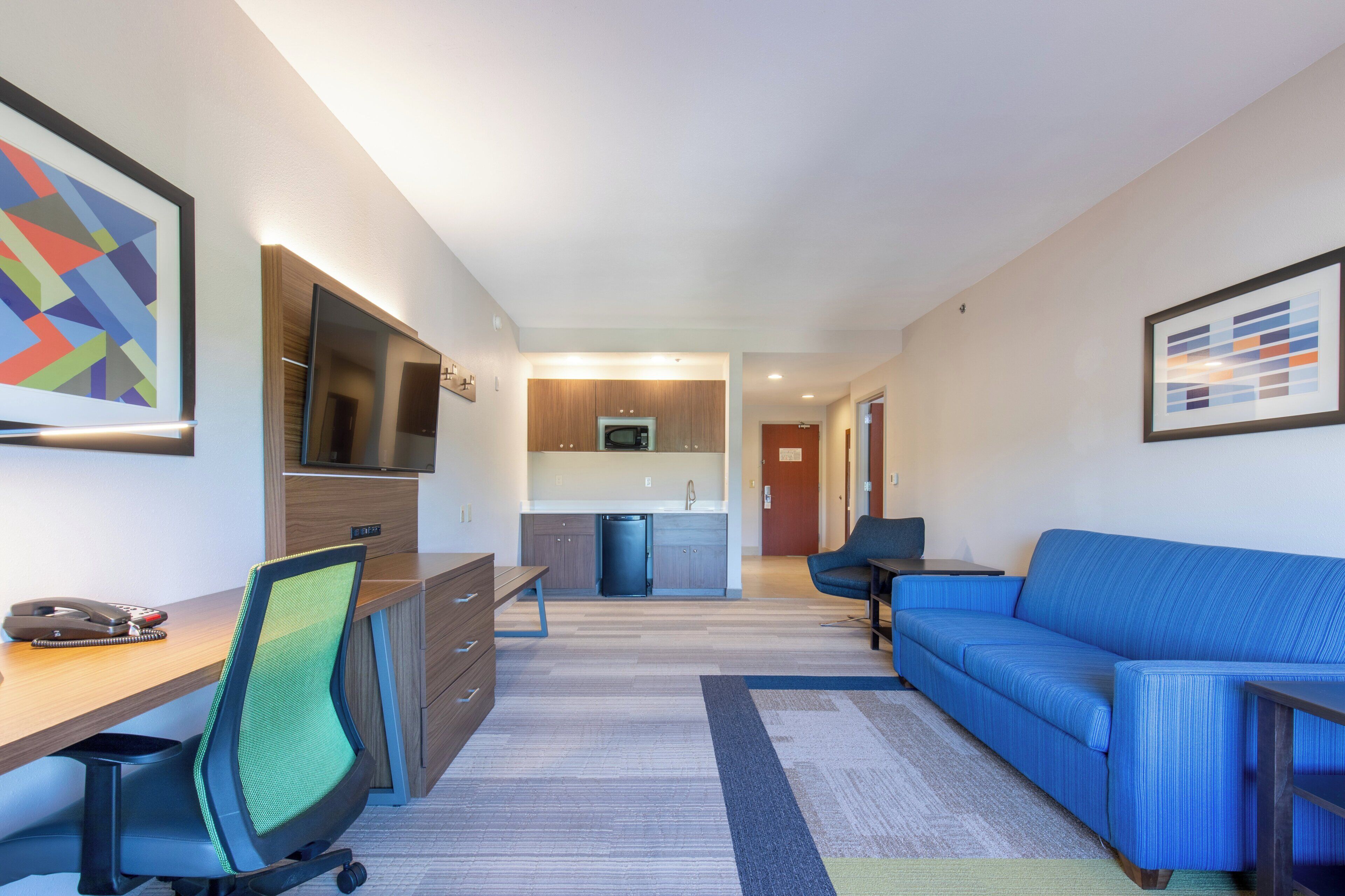 A modern living room with a blue sofa, a desk with a chair, a television, and a kitchen area in the background.