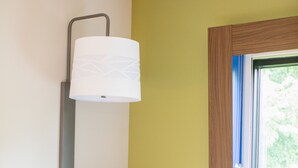 A corner room with a grey upholstered chair, a wall-mounted lamp, and a window with a view of greenery. - Holiday Inn Express Johnson City by IHG (Johnson City)