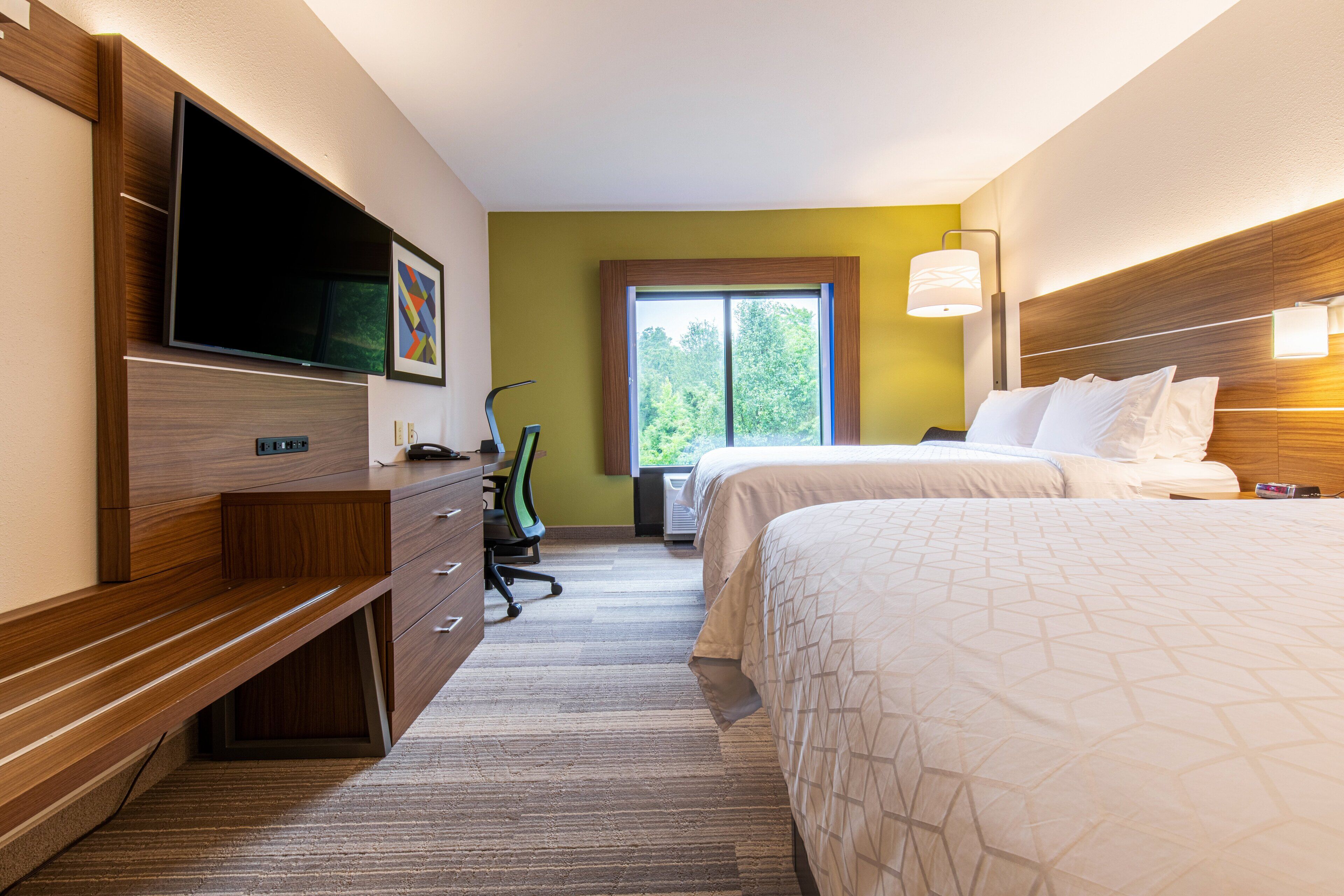 A hotel room with a bed, a desk with a chair, a television, and a window with a view of trees.