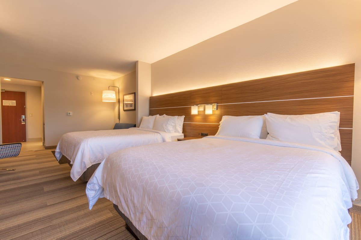 Habitación de hotel con dos camas, cabecero de madera, luces fijadas a la pared y suelo cubierto de alfombra.