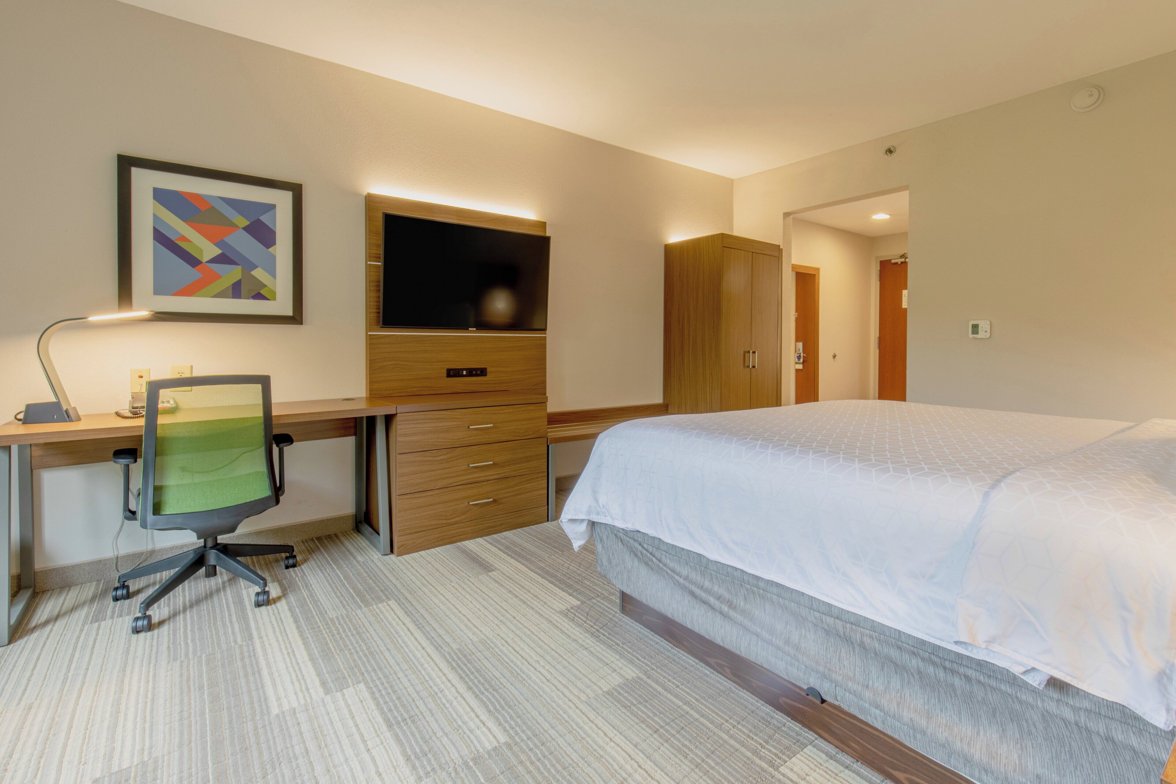Une chambre d’hôtel équipée d’un lit, d’un bureau, d’une chaise, d’une télévision, d’une armoire et d’œuvres d’art accrochées au mur.