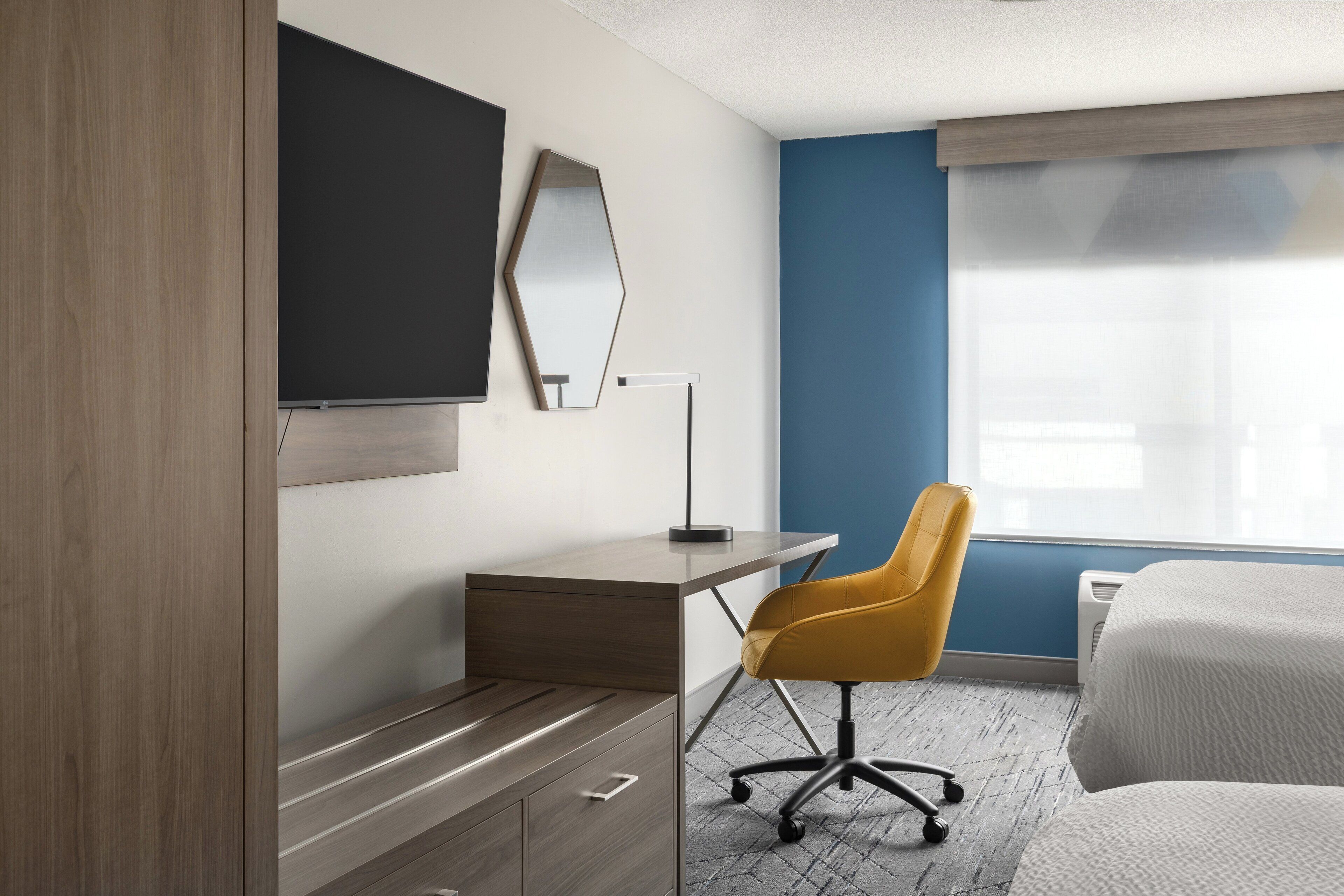 Une chambre d’hôtel avec un bureau en bois, une chaise jaune, un lit, un grand miroir et une fenêtre avec des stores.