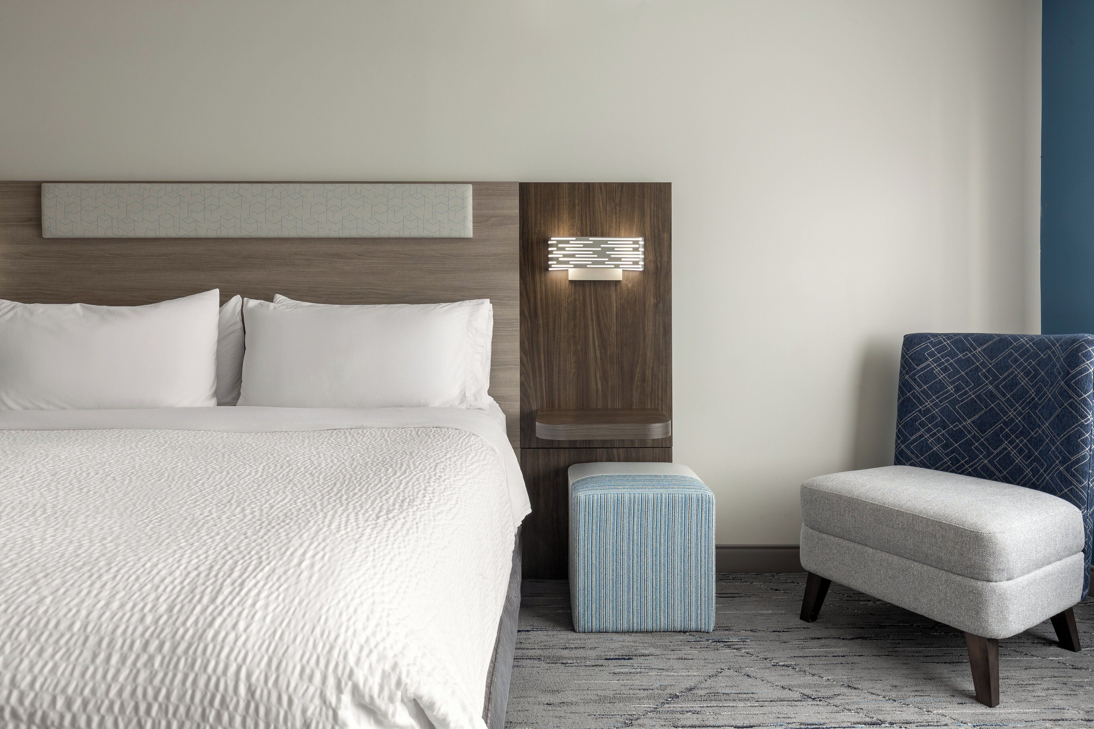 A modern hotel room with a large bed, a small ottoman, a chair, and a wall-mounted light fixture.