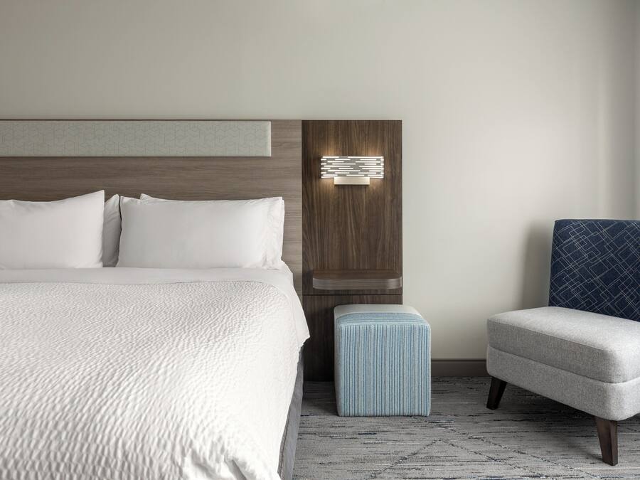 A modern hotel room with a large bed, a small ottoman, a chair, and a wall-mounted light fixture.