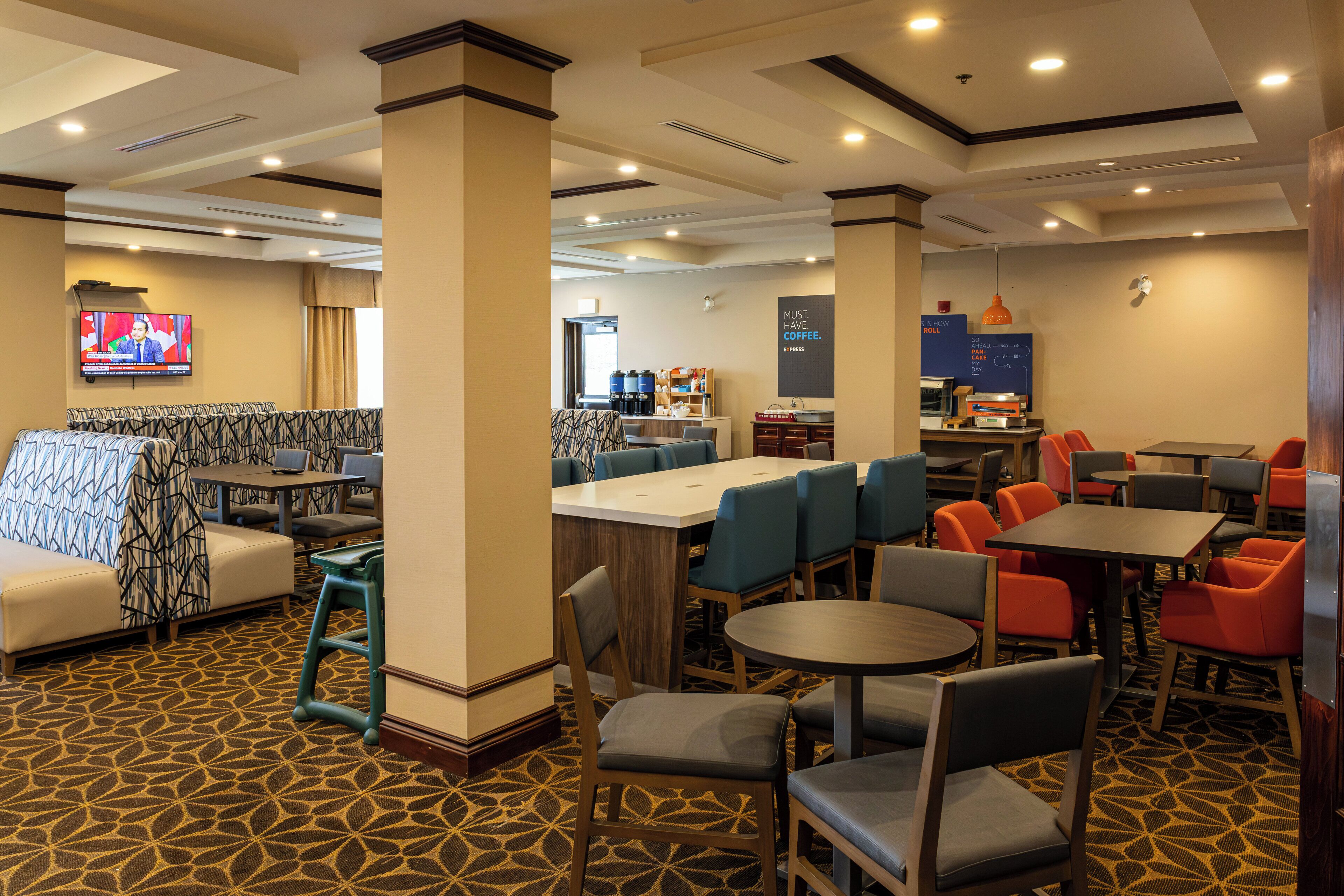 A hotel lobby with a patterned carpet, multiple seating areas including a bar and dining tables, and a television mounted on the wall.