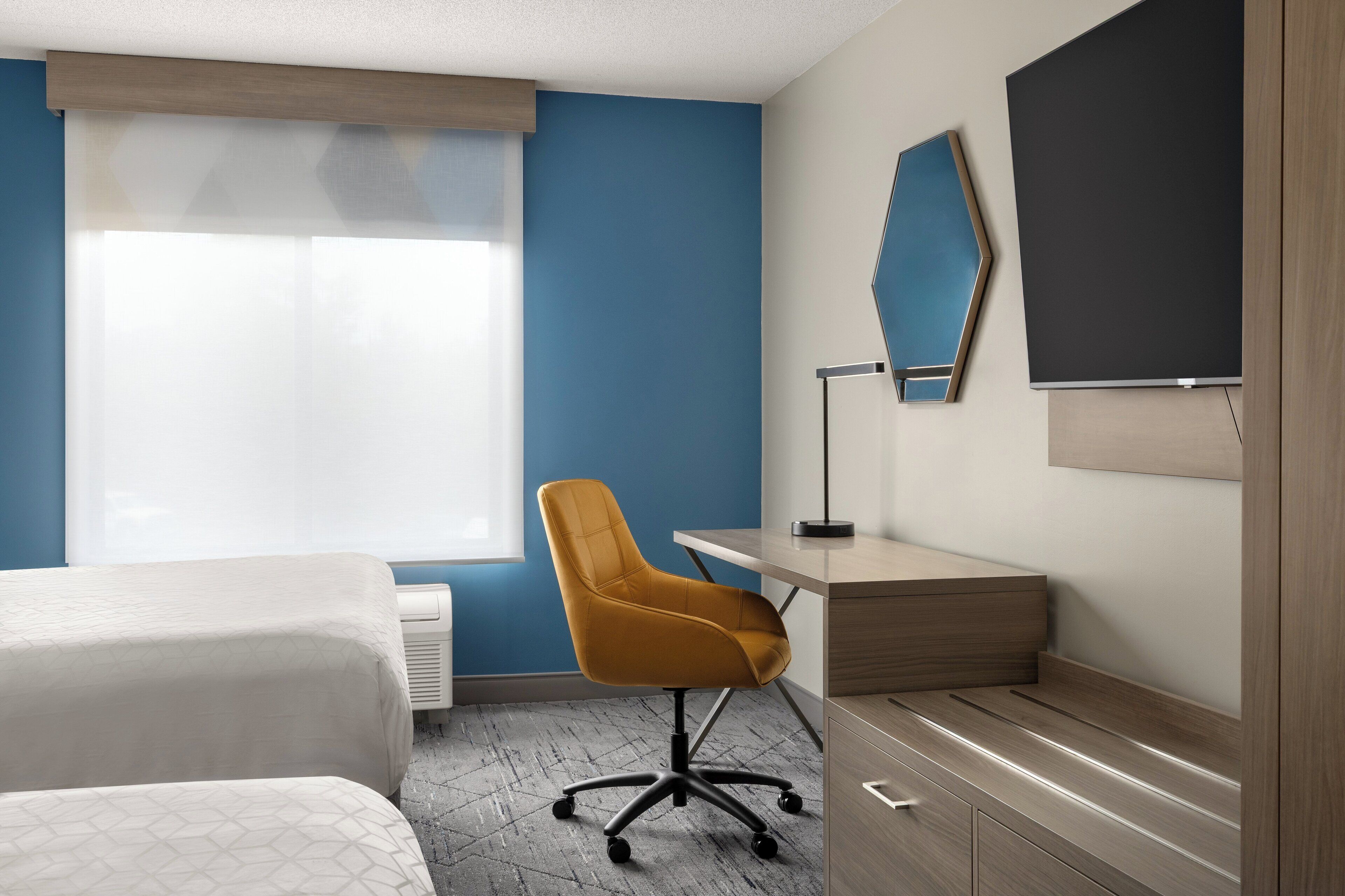 Habitación de hotel con cama, escritorio, silla, televisor y ventana con persianas.