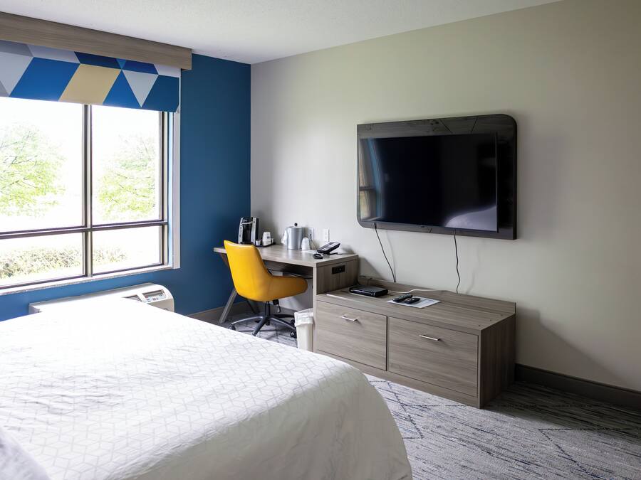 A hotel room with a bed, a desk with a chair, a television, and a window with blinds.