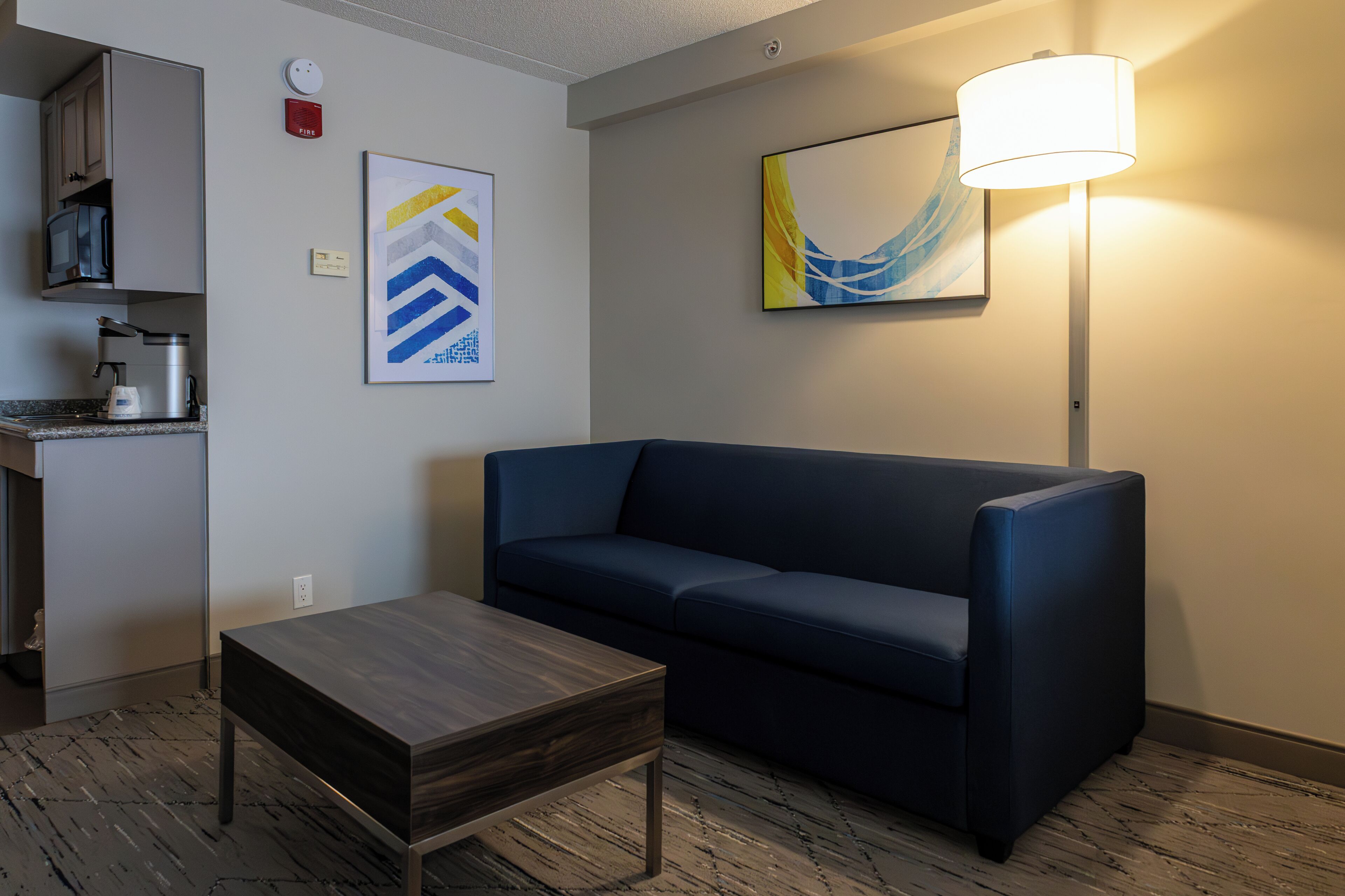 Een moderne hotelkamer met een donkerblauwe bank, een klein houten tafeltje, een lamp, een magnetron en een koffiezetapparaat.