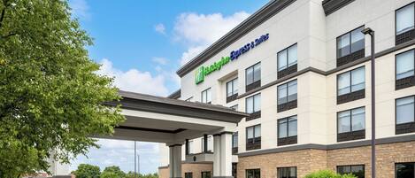 Un edificio de Holiday Inn Express & Suites de diseño moderno, con entrada cubierta y zona ajardinada con árboles y arbustos.