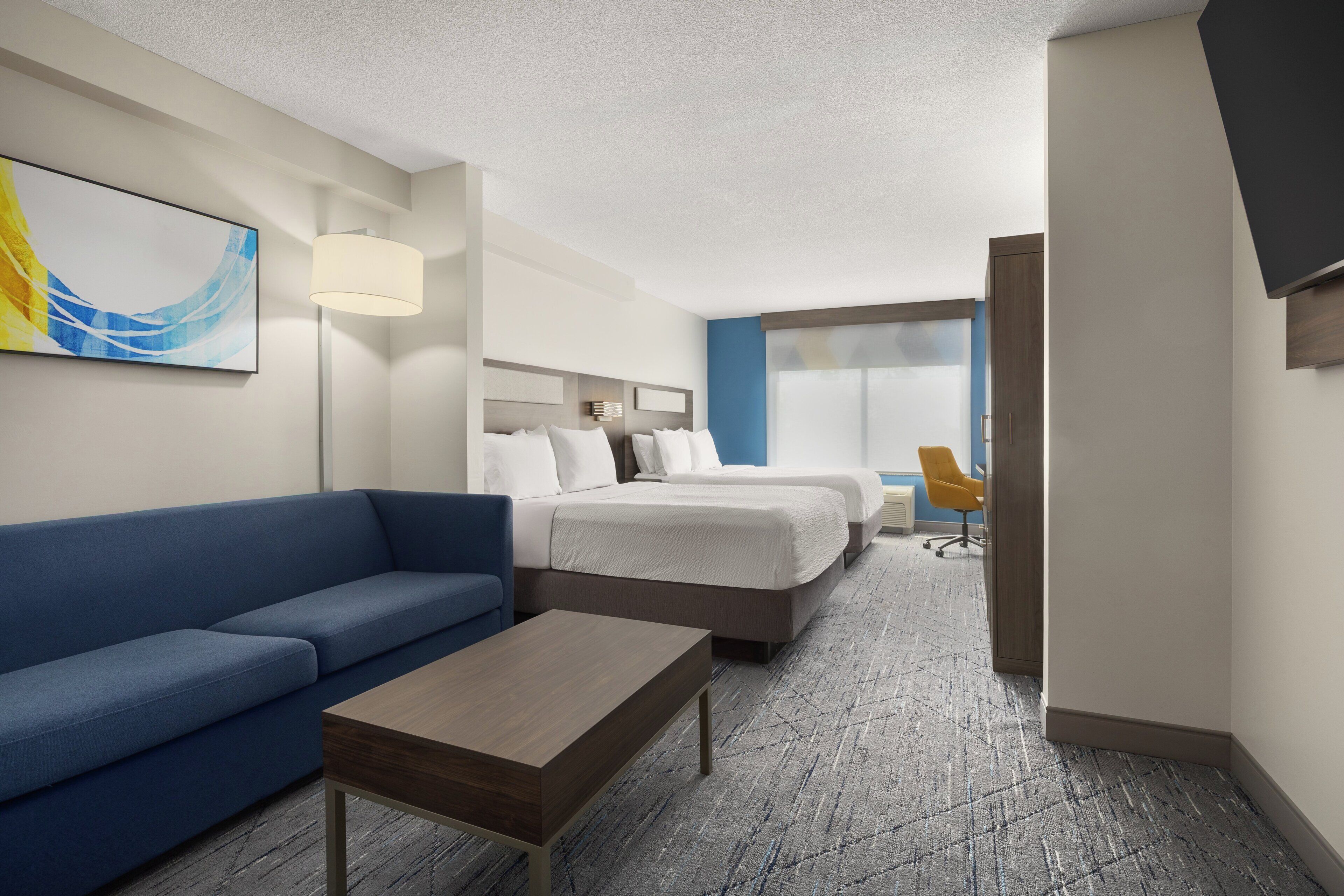 Habitación de hotel con un sofá azul, una mesa de centro de madera, dos camas con ropa blanca, un escritorio con una silla y un cuadro abstracto grande en la pared.