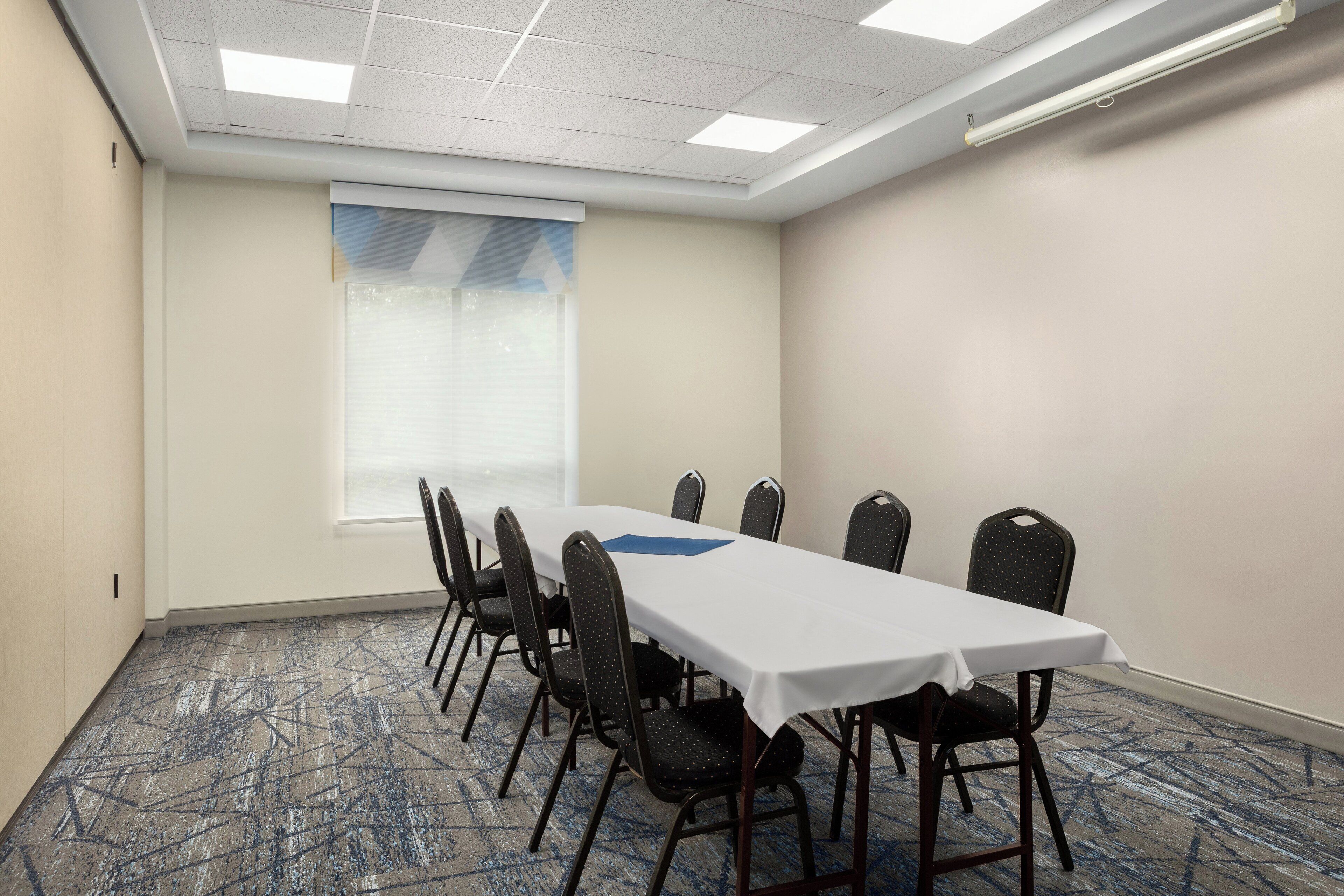 Una sala de conferencias con una larga mesa, sillas, una ventana con persianas y suelo alfombrado.