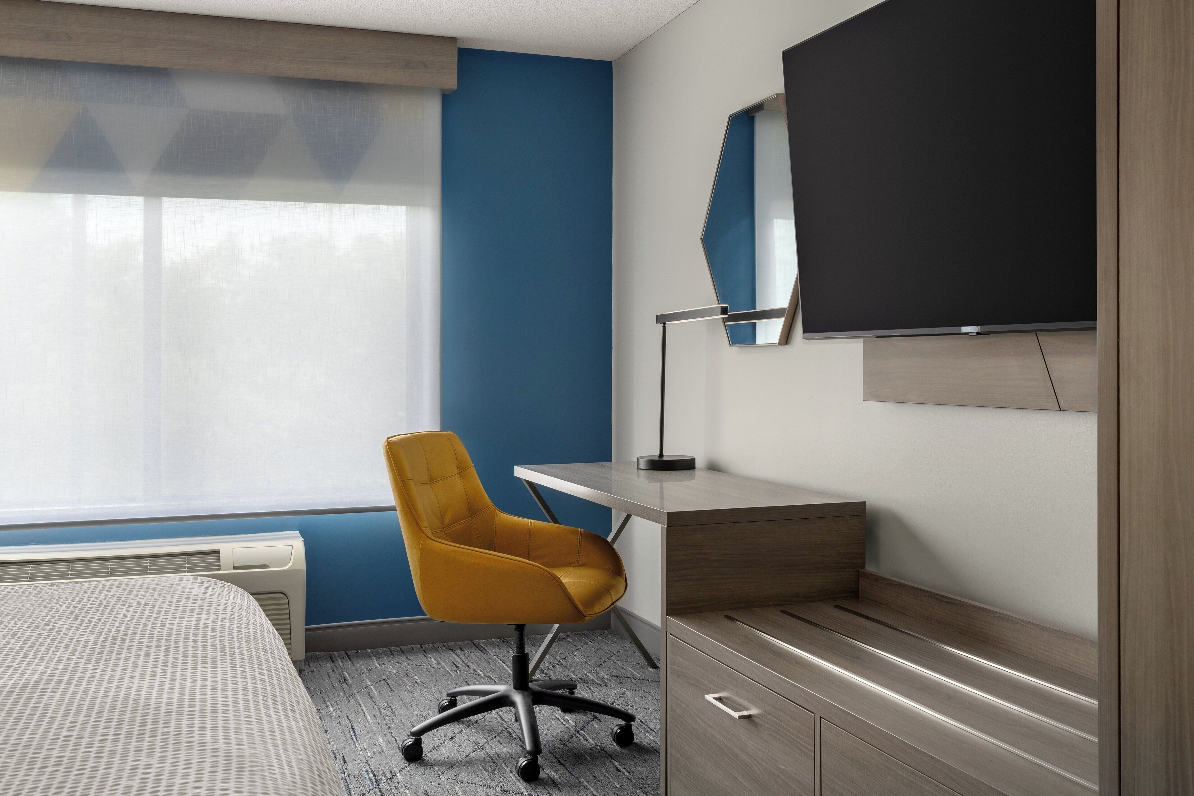 Quarto de hotel com cama, escrivaninha e cadeira, televisão de tela plana e janela com persianas.
