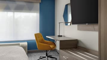 Quarto de hotel com cama, escrivaninha e cadeira, televisão de tela plana e janela com persianas.