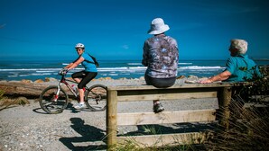 A wooden bench overlooking a beach with a cyclist and an elderly person sitting on it. - Charles Court Motel (Greymouth)