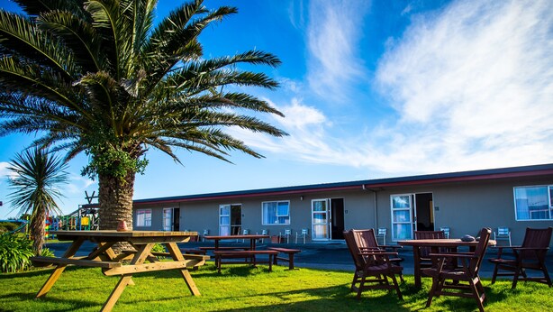 A building with numbered rooms, outdoor seating, and a palm tree. - Charles Court Motel (Greymouth)