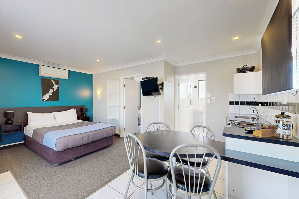 Une chambre d’hôtel compacte comprenant un lit, une petite table à manger et des chaises. Elle dispose également d’une kitchenette avec un évier et une cuisinière.