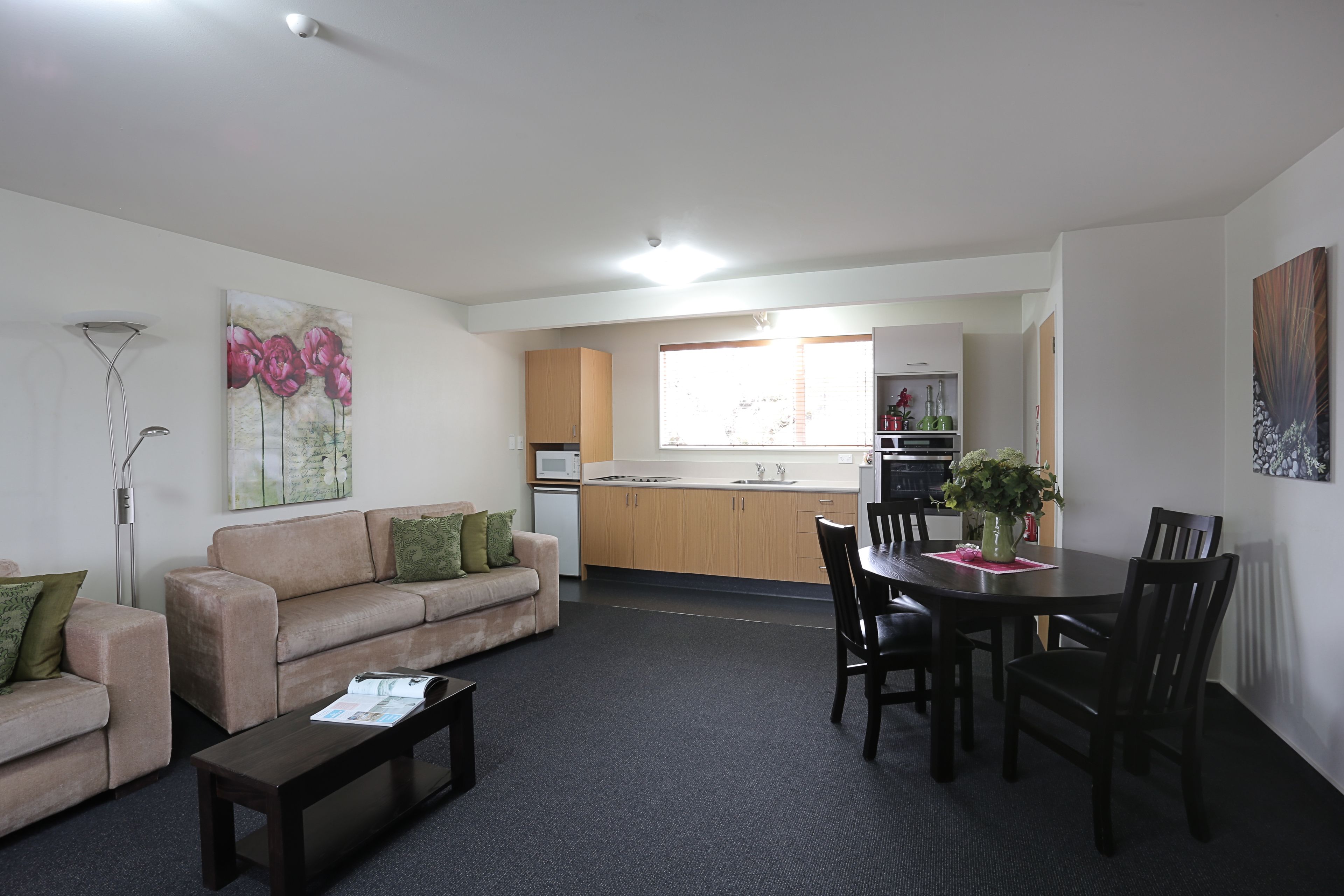 A modern living room with a sofa, coffee table, and dining area with chairs and a table. A kitchen with cabinets and appliances is visible in the background.
