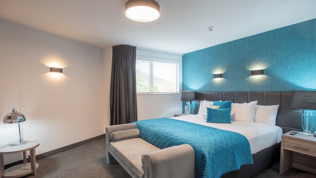 A modern hotel room with a large bed, a bedside table, a lamp, and a window with curtains. - Homestead Villa Motel (Invercargill)