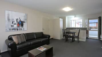 A modern living room with a black leather sofa, a coffee table, and a dining area with a table and chairs. A cityscape painting is on the wall, and a kitchen is visible through an open door.