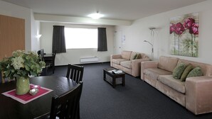 A living room with a dining table, two sofas, a coffee table, and a large window with curtains. - Homestead Villa Motel (Invercargill)
