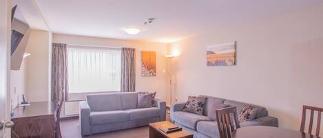 A modern living room with a grey sofa, a wooden coffee table, and a dining table with chairs. There is a painting on the wall and a floor lamp.