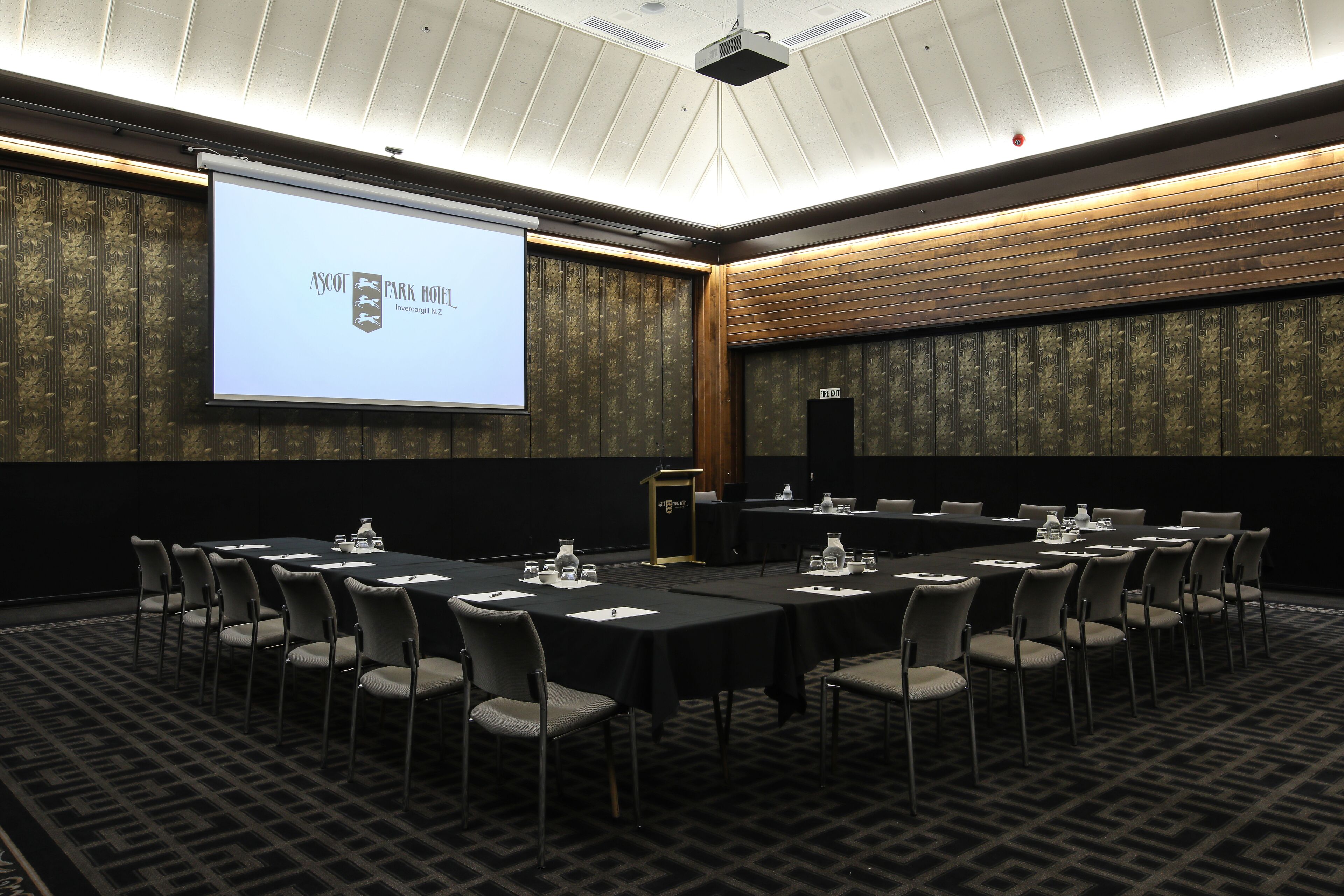 Un salón de conferencias con una pantalla grande mostrando una presentación, varias mesas con sillas y un atril.