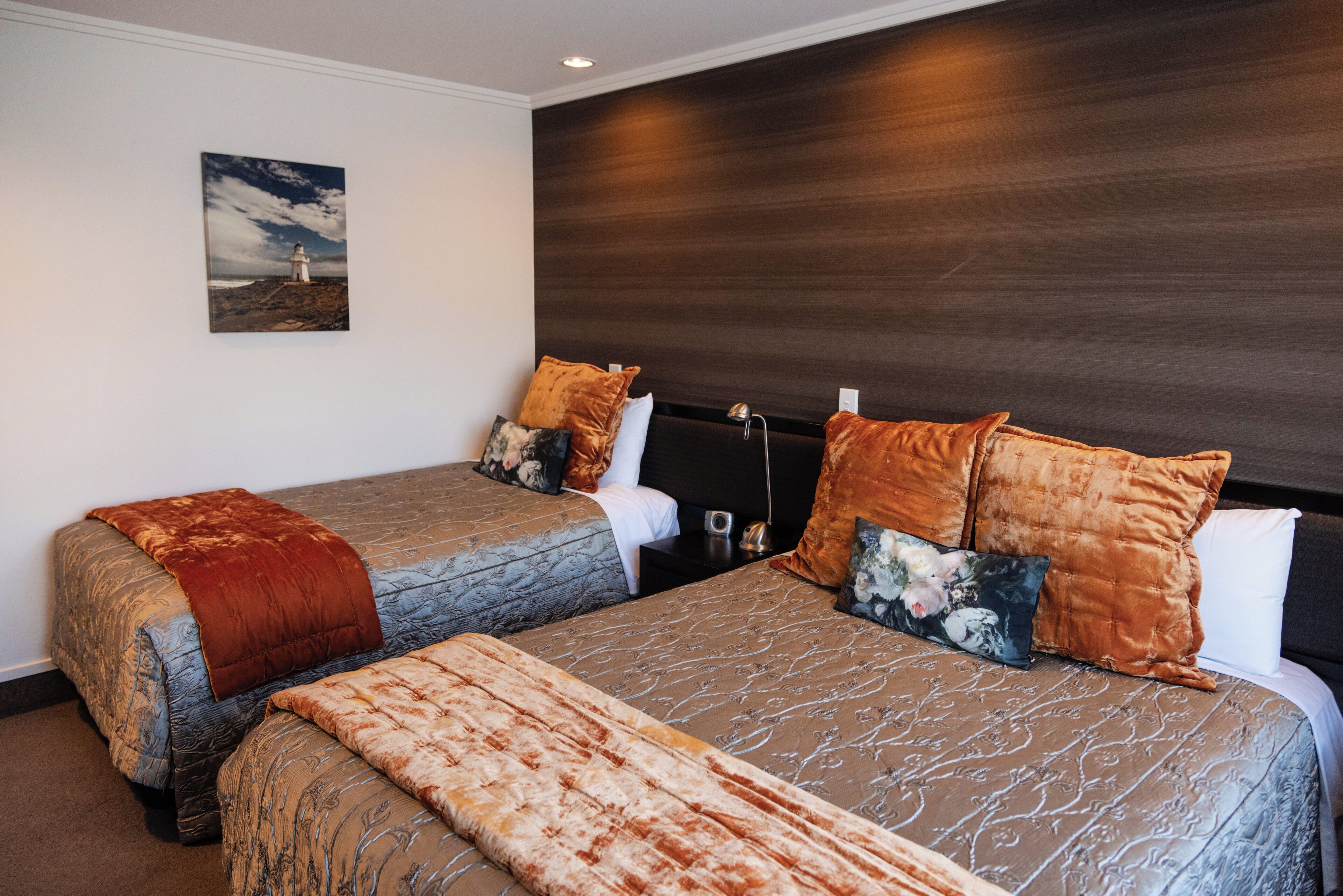 Habitación de hotel con dos camas, cabecera de madera oscura, un cuadro de un faro y una colcha de edredón estampada en tonos marrones y azules.