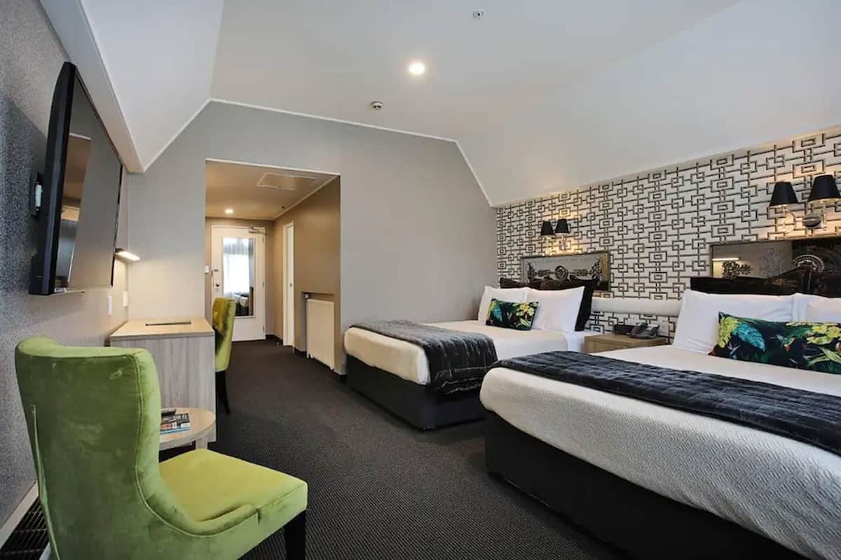A hotel room with two beds, a green chair, a desk, and a patterned wall.