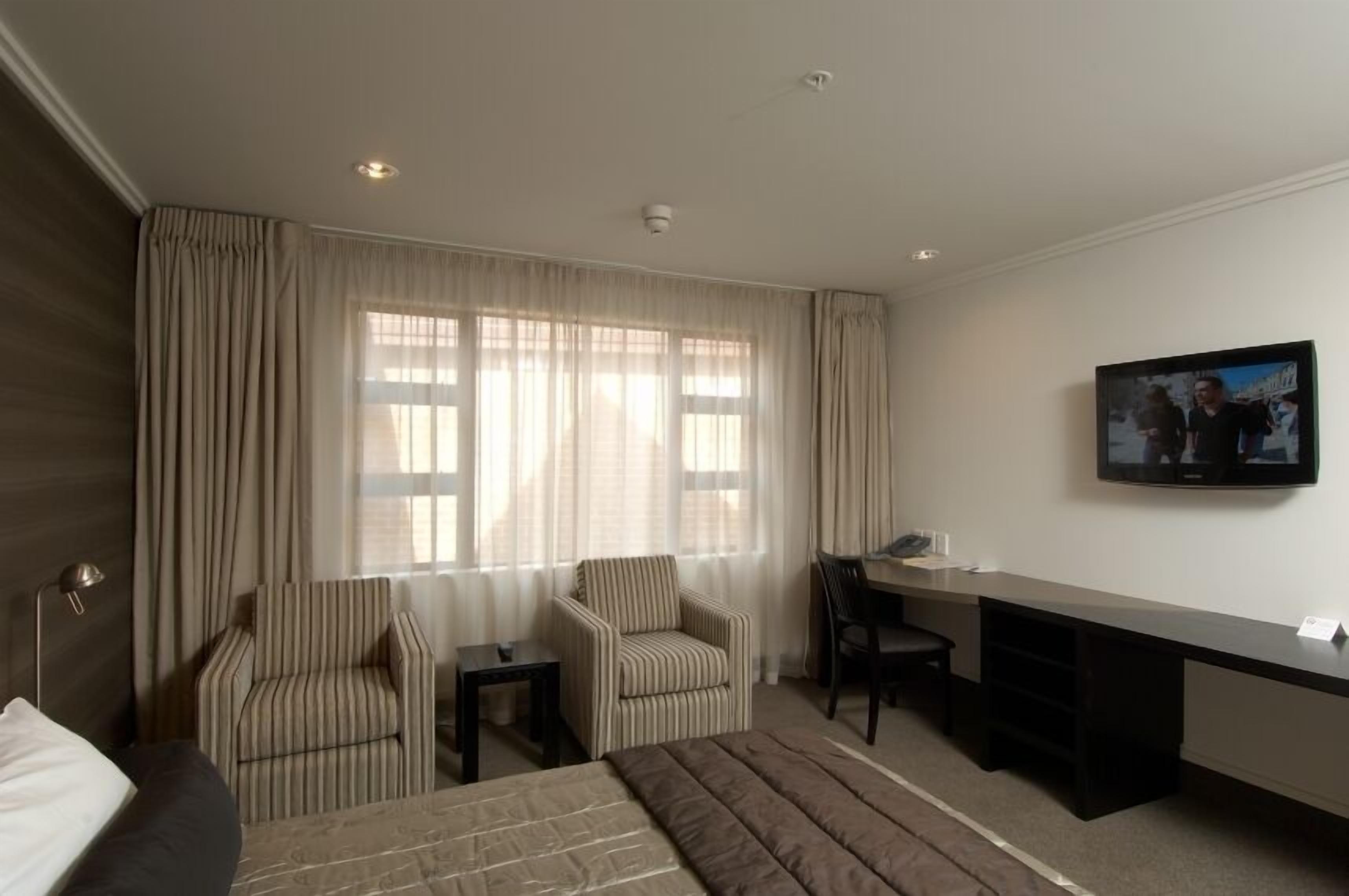A hotel room with a bed, two striped armchairs, a desk, and a television on the wall.