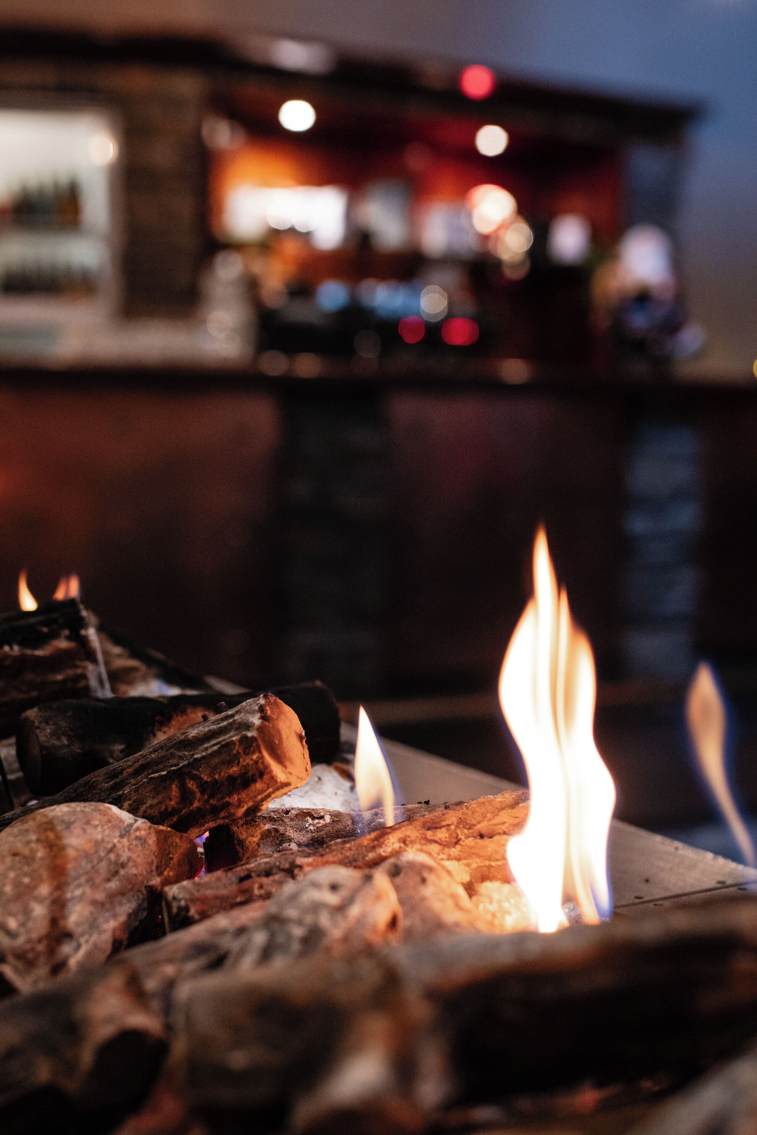 A close-up of a fireplace with burning logs and flames, with a blurred background suggesting a bar area.