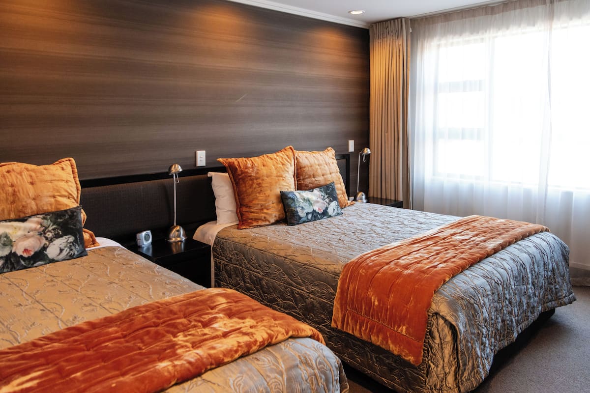 A hotel room with two beds, each with a duvet and pillows, a bedside table with a lamp, and a window with curtains.