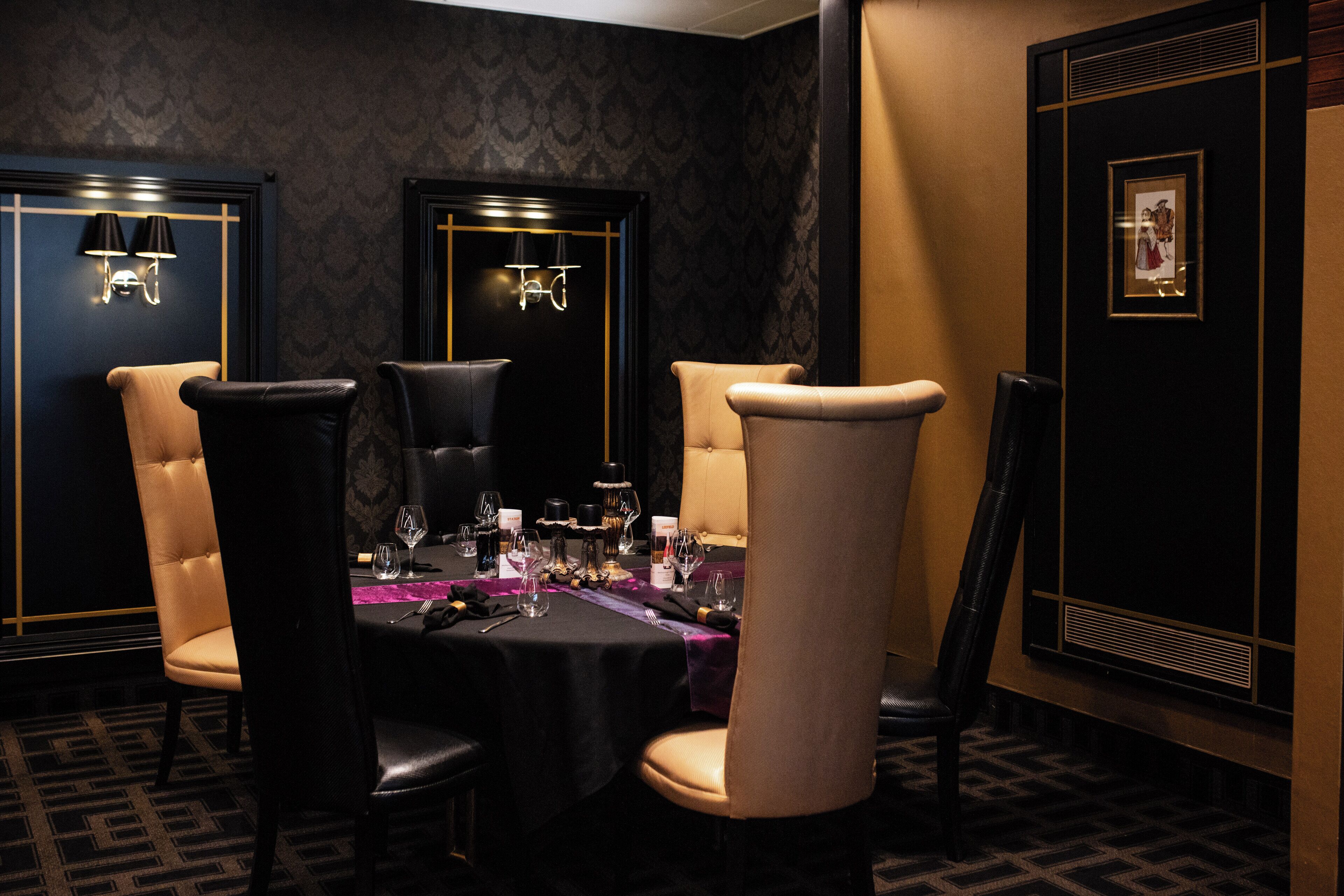 A dining area with a table set for four, surrounded by high-backed chairs, with wall sconces and a framed picture on the wall.