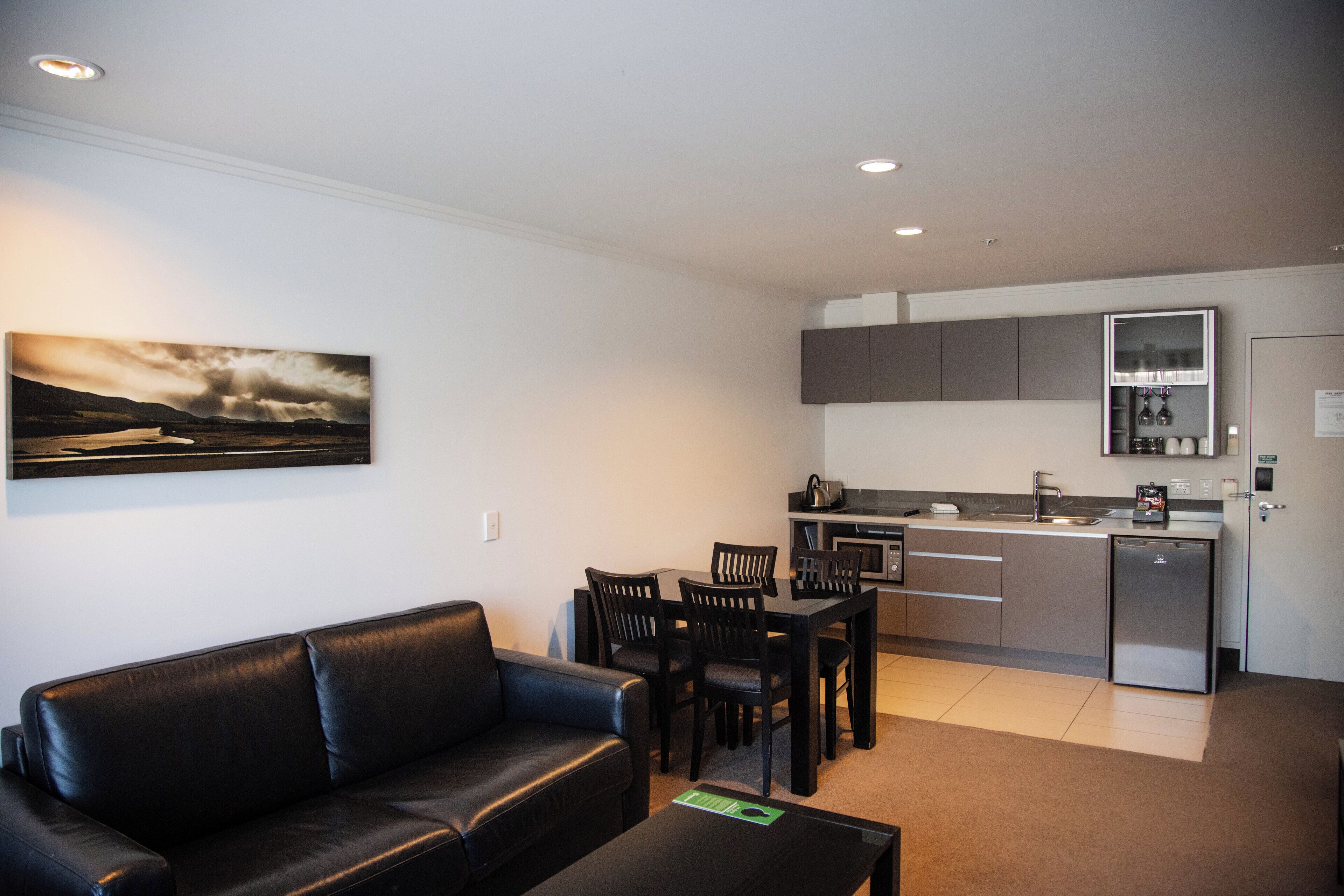 Un apartamento moderno con sala de estar que incluye un sofá de cuero negro, una mesa de comedor con sillas, una cocina con armarios grises y un refrigerador.