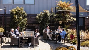 An outdoor seating area with wicker furniture and a table, surrounded by plants and a building with windows. - Ascot Park Hotel (Invercargill)
