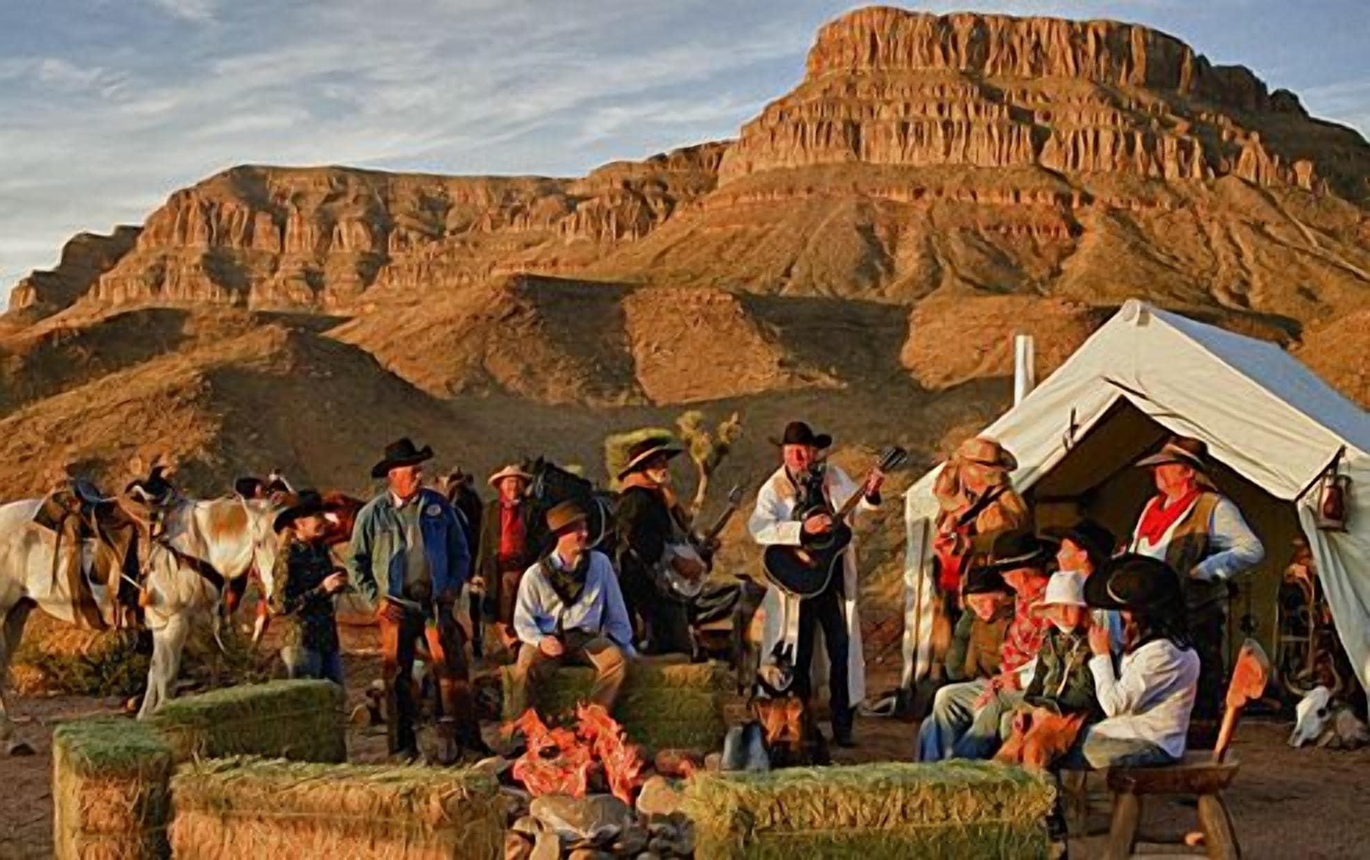 A group of people gathered around hay bales in a desert landscape with a tent and a large rock formation in the background.